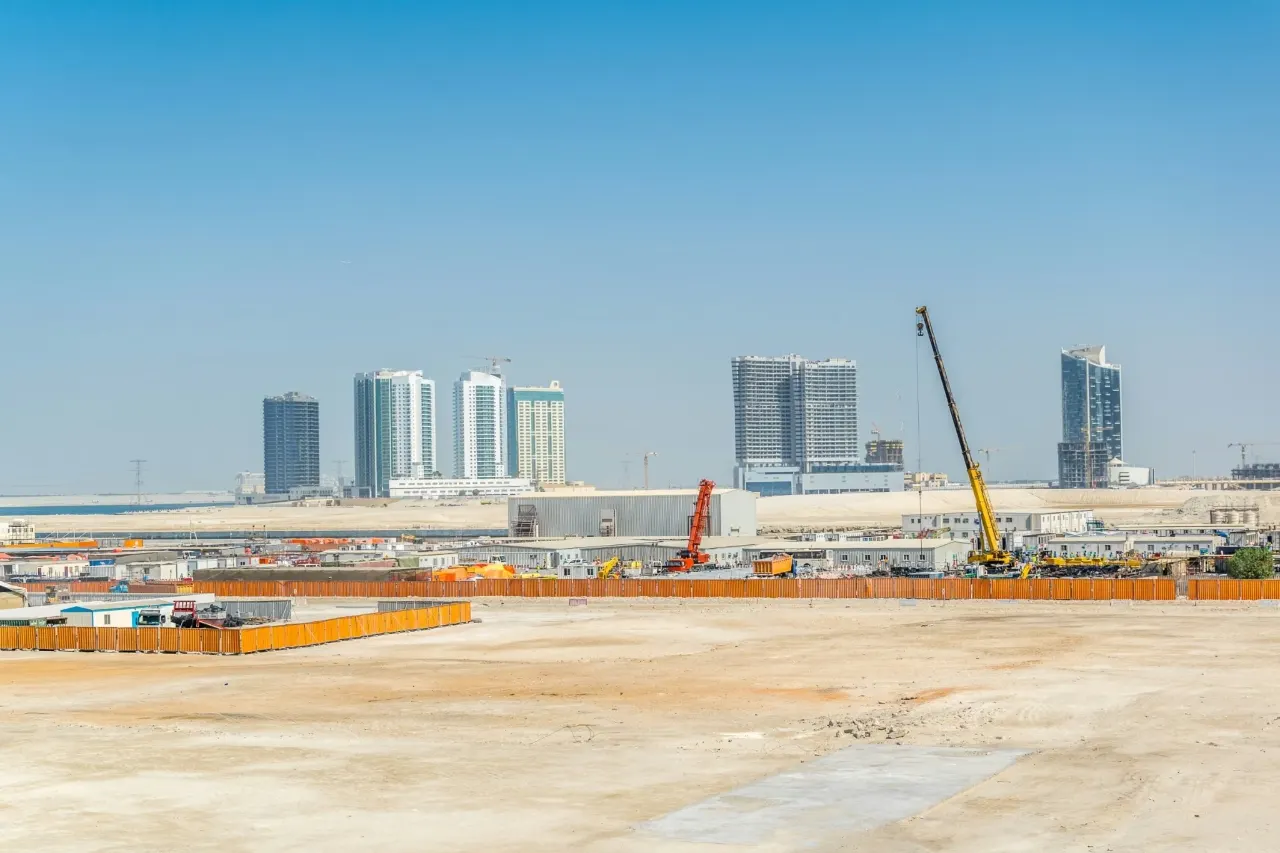 A cleared, fenced construction site with mobile cranes and site cabins, and towers on the skyline beyond