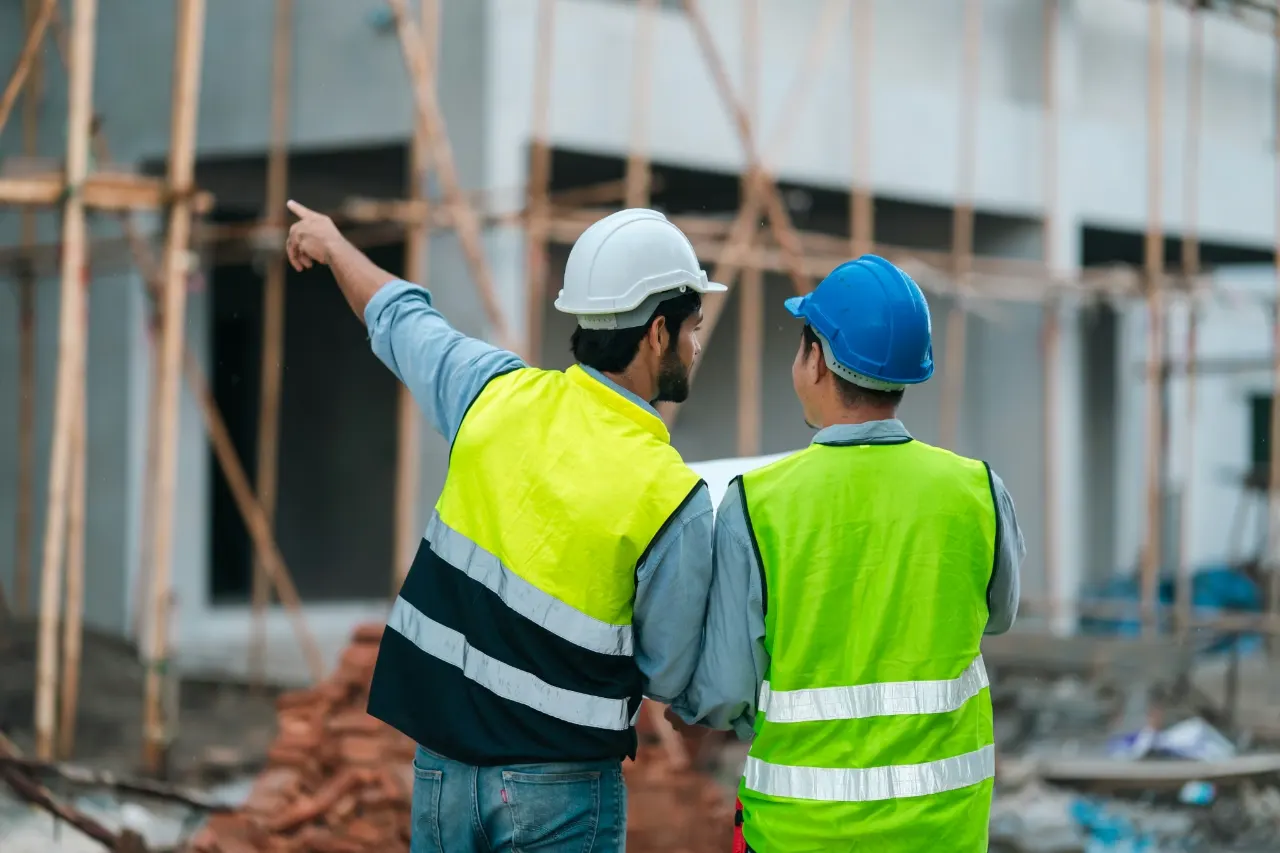 Two site professionals in hard hats and high-visibility vests reviewing a building under construction