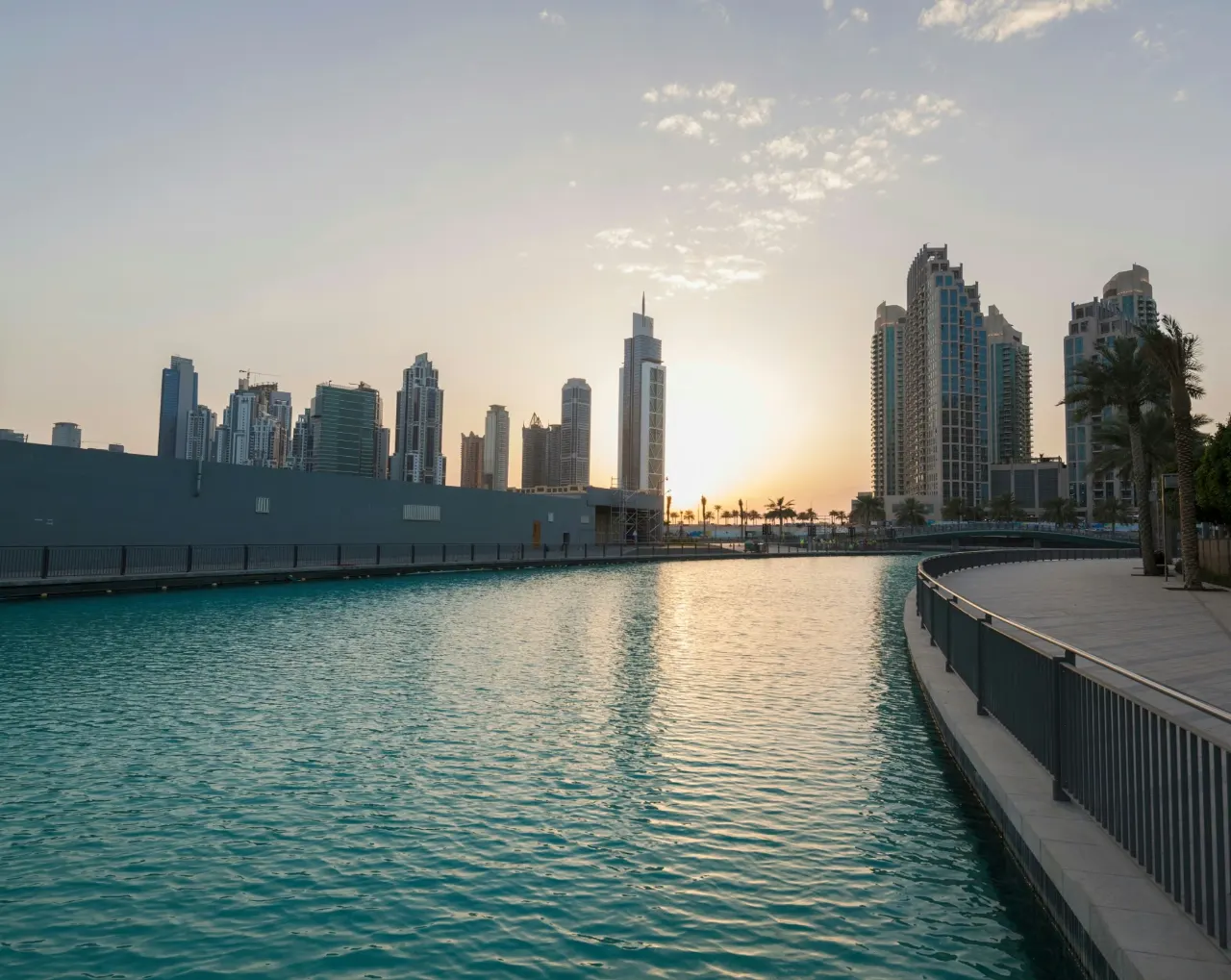 The sun setting behind high-rise towers, seen across a calm turquoise canal with a railed waterside walkway