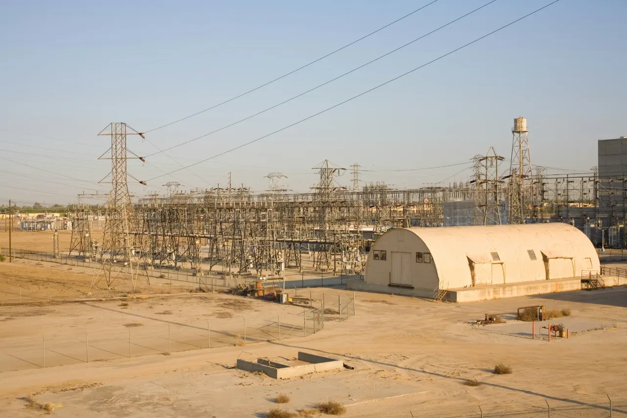 An electrical substation and transmission towers on open, dry ground beside a curved-roof service building