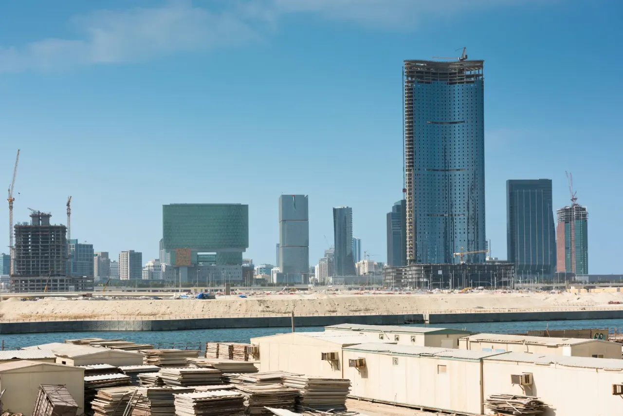 Site cabins and stacked formwork on reclaimed ground, with a new district of towers and roads under construction across the water