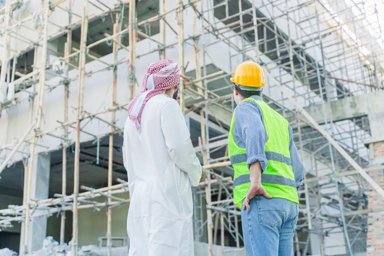 A man in kandura and a site engineer in a hard hat and vest review a building under construction behind scaffolding