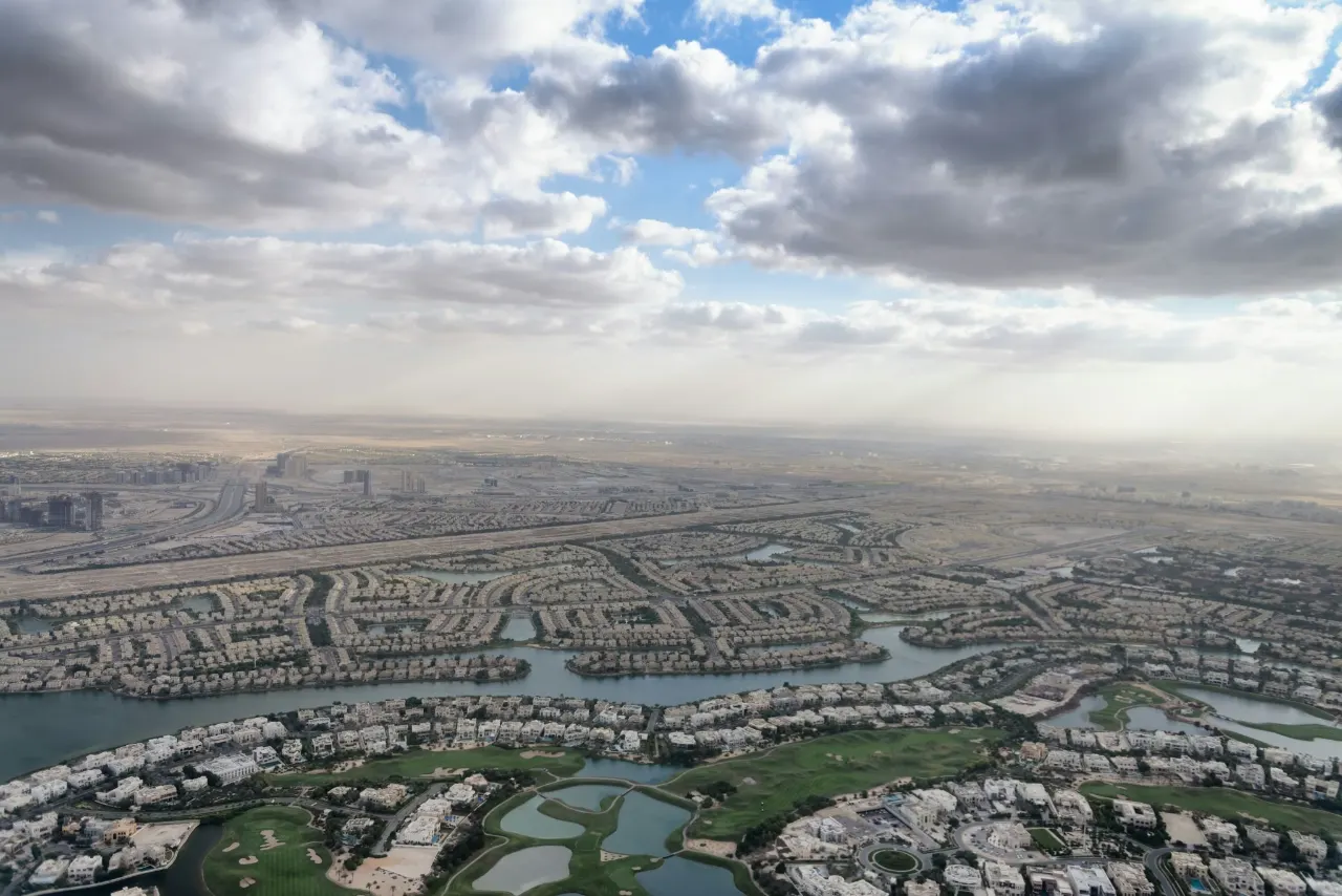 Aerial view over the edge of Dubai: residential districts, waterways and road corridors meeting open desert under broken cloud