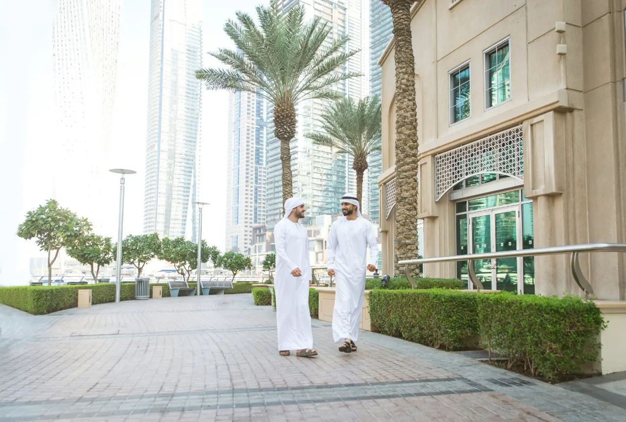 Two men in kandura talking as they walk along a paved, landscaped promenade between residential buildings