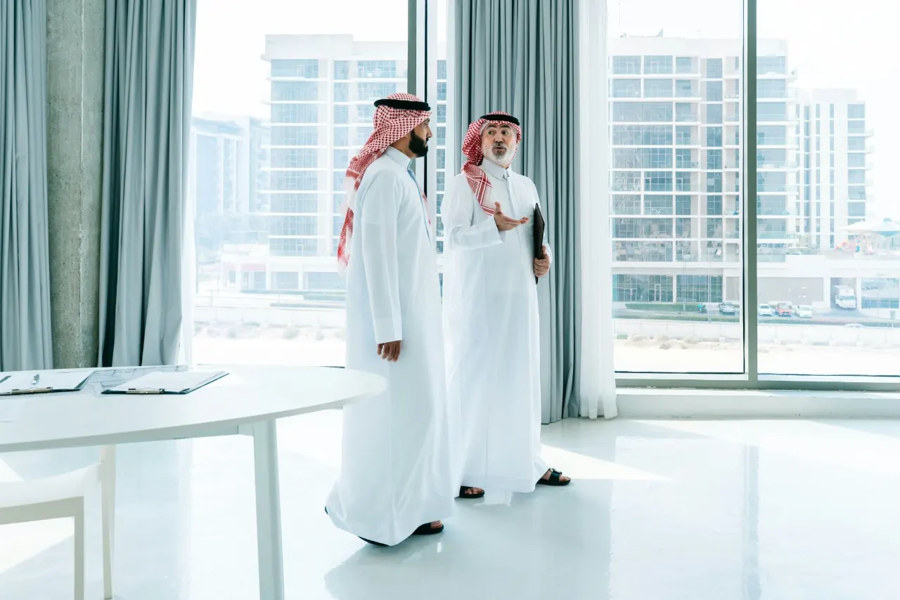 Two men in white kandura and red-checked headscarves talking by floor-to-ceiling windows in a bright, empty office; one holds a folder