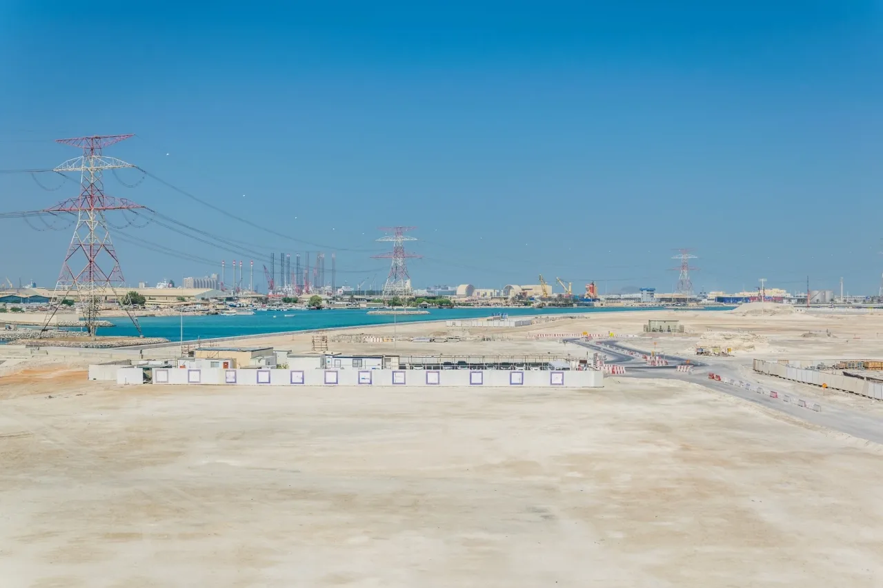 Cleared waterfront land with a site compound, access road and power lines, with the port and industry beyond