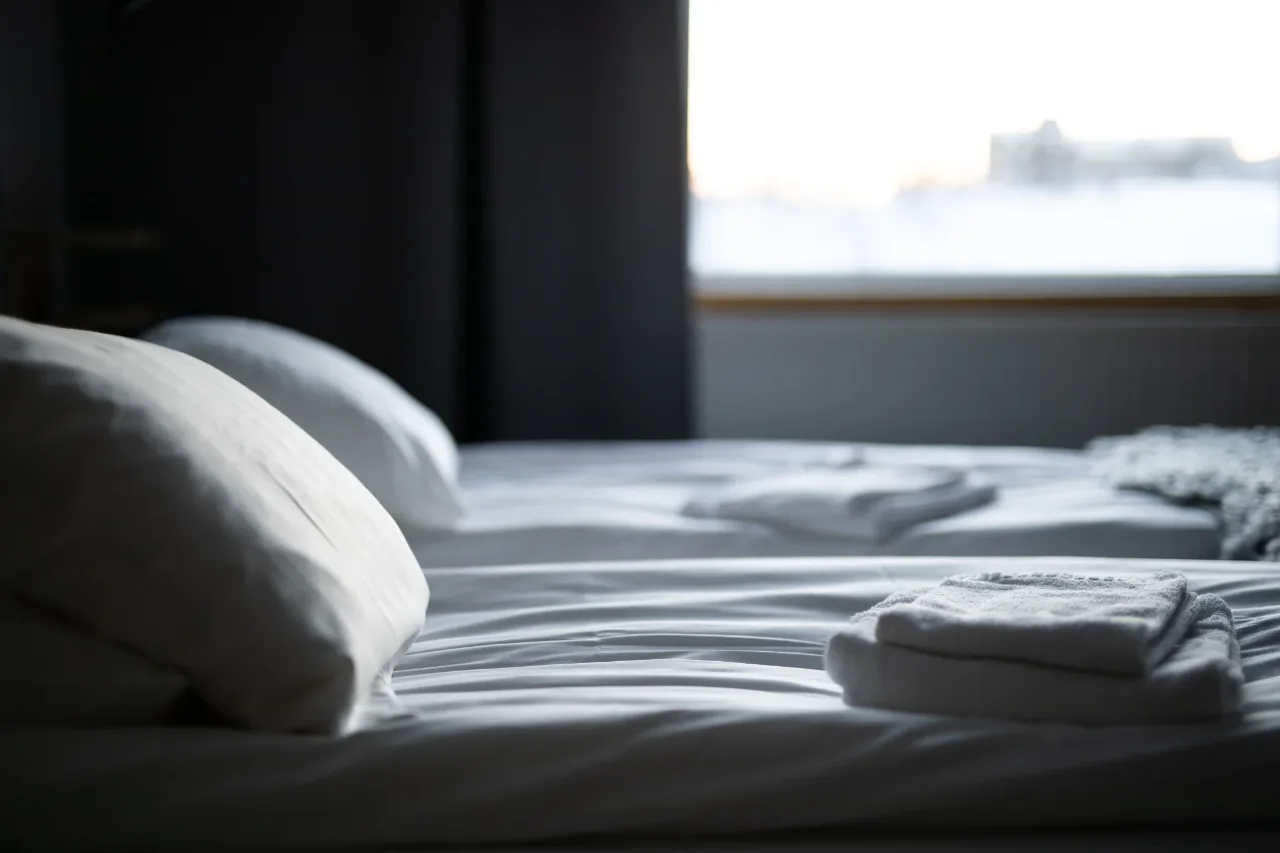 A made bed with fresh linen and folded towels beside a window, in quiet natural light