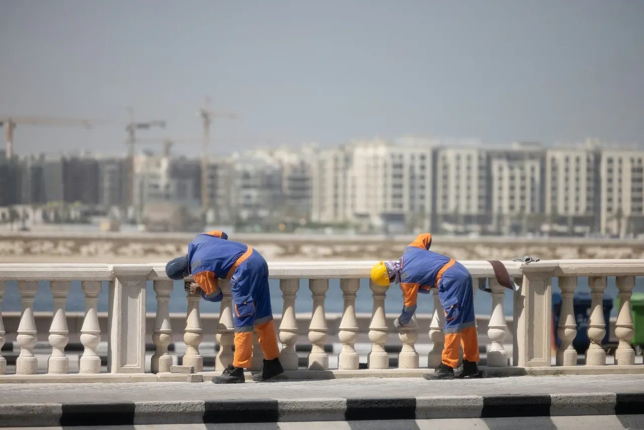 Two maintenance workers in overalls finishing a stone balustrade on a waterfront promenade, with residential blocks and cranes beyond